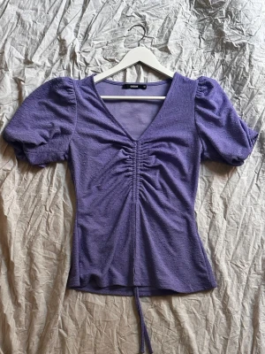 Lila topp med puffärm och snörning - Superfin lila topp från Bik Bok med v-ringning, puffärmar och snörning framtill som ger en snygg rynkad effekt. Toppen har en figurnära passform och är gjord i ett mjukt, lätt strukturerat material.