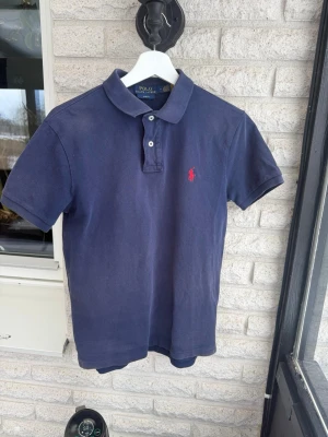 Marinblå pikétröja från Polo Ralph Lauren - Klassisk marinblå pikétröja från Polo Ralph Lauren med korta ärmar och krage. Tröjan har två knappar framtill och den ikoniska röda logotypen broderad på bröstet. Perfekt till jeans eller shorts för en avslappnad men snygg stil.