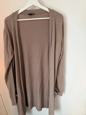 Beige lång kofta från new yorker  - Lång, öppen beige kofta från new yorker med långa ärmar. Tillverkad i ett mjukt och tunt material som ger en avslappnad look. Perfekt att slänga över en t-shirt eller topp för extra lager. Enkel och stilren design med två fickor. 