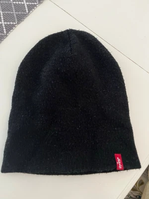 Svart mössa från Levi's - Säljer en klassisk svart mössa från Levi's med den ikoniska röda loggan på kanten. Mössan är ribbstickad och kan bäras både med eller utan uppvikt kant. Perfekt för att hålla värmen och ge en clean look.