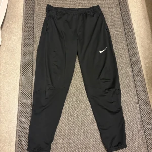 Svarta träningsbyxor från Nike - Säljer ett par svarta träningsbyxor från Nike med vit logga på vänster lår. Byxorna har dragkedjor vid fickorna och resår i midjan. Perfekta för gymmet eller löprundan. Tillverkade i ett lätt och stretchigt material.