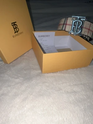 Burberry skärp - Säljer ett ikoniskt Burberry skärp med klassiskt rutigt mönster i vitt, svart och rött. Skärpet har ett stort silvrigt BB-spänne framtill och är tillverkat i skinn. Perfekt accessoar för att lyfta vilken outfit som helst.