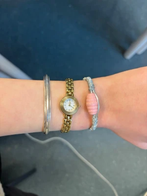 Silvrigt armband med rosa pärla - Unikt armband i silverfärgad metall med en stor, räfflad rosa pärla i mitten. Armbandet har en öppen design med tvinnad metalltråd runt bandet och dekorativa svarta detaljer vid ändarna. Perfekt för dig som gillar handgjorda accessoarer med personlig touch.