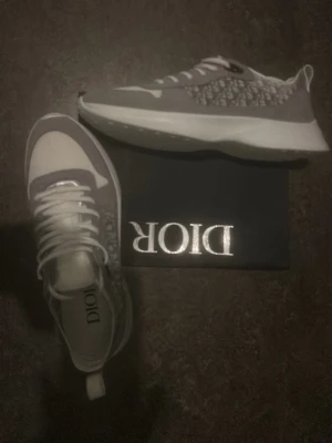 Gråa Dior sneakers med mönster - Säljer ett par gråa Dior sneakers med vitt och grått mönster på sidan och vita snören. Skorna har en chunky vit sula och klassisk Dior-logga på innersulan. Perfekta för dig som vill ha något stilrent men ändå sticker ut. Passar även mycket bra för storlek 44 och 44,5