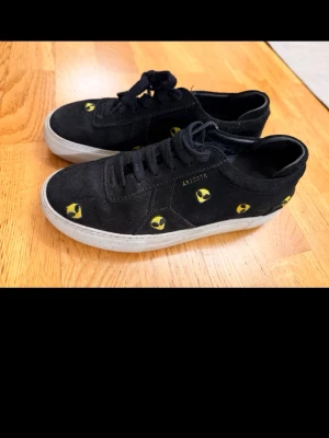 Svarta sneakers från Axel Arigato - Svarta sneakers i mocka från Axel Arigato med broderade gula smileys över hela skon.  Märkeslogga i guld på sidan. Perfekta för dig som vill ha något unikt och trendigt.