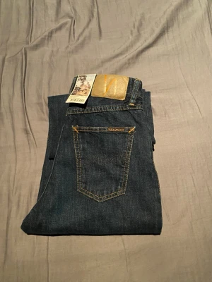 Mörkblå jeans från Nudie Jeans - Säljer ett par mörkblå jeans från Nudie Jeans med klassisk femficksmodell och kontrastsömmar i gult. Jeansen har en rak passform och är tillverkade i slitstark denim med Nudie-loggan på bakfickan och patch i läder baktill pris kan diskuteras.