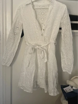 Vit broderad kort klänning med knyt - Söt vit kort klänning med broderade blommor, knappar framtill och knytband i midjan. Klänningen har lång ärm och är tillverkad i luftig bomull, perfekt för varma dagar. Ger en romantisk och fräsch känsla.