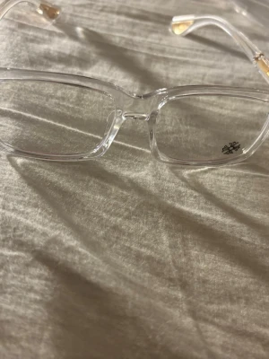 Chrome heart glasögon  - Säljer ett par rektangulära solglasögon från Chrome Hearts med transparent båge och snygga gulddetaljer på skalmarna. Glasögonen har en unik logga på linsen och ger en clean och modern vibe.