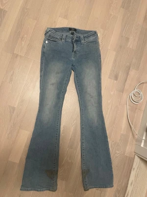 Blå bootcut jeans från True Religion - Säljer ett par blå bootcut jeans från True Religion med klassisk femficksdesign och snygga detaljer på bakfickorna. Jeansen har en lätt utsvängd passform och är tillverkade i bomull med stretch för extra komfort. Perfekt för dig som gillar en avslappnad och trendig stil.