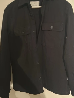 Svart overshirt från Jack & Jones - Stilren svart overshirt från Jack & Jones med klassisk krage och två bröstfickor med knappstängning. Skjortan har raka ärmar och knappar framtill. Perfekt att bära öppen eller stängd för en avslappnad look.