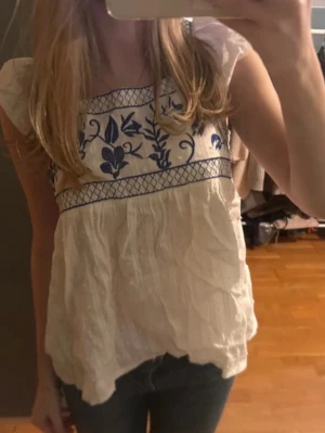 Vit blus med blå broderier - Söt vit blus med blå broderade blommor och grafiskt mönster upptill. Blusen har korta ärmar och luftig passform, perfekt för varma dagar. Tillverkad i lätt bomullstyg och har en avslappnad, flowy siluett.