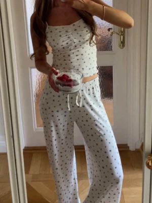 Vit pyjamas sett lager 157 blåbär - Säljer ett vitt loungewear-set med linne och byxor, båda med små svartblå blommor. Linnet har knappar framtill och byxorna har dragsko i midjan för skön passform. Perfekt för chill dagar hemma.