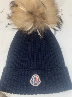  Moncler mössa med pälsboll äkta - Snygg mörkblå ribbstickad mössa från Moncler med stor beige pälsboll på toppen och klassisk Moncler-logga framtill. Perfekt accessoar för kalla dagar och ger en lyxig touch till din outfit.