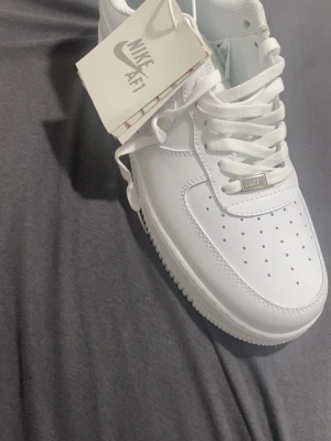 Nike Air Force 1 vita sneakers - Klassiska Nike Air Force 1 sneakers i helvitt läder med perforerad tåbox, vita skosnören och diskret Nike-logga. Tidlös design med låg profil och robust sula. Perfekta för dig som gillar clean och stilren look.