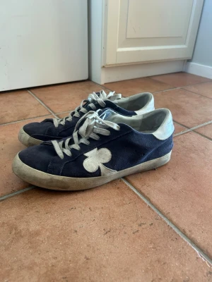 Mörkblå sneakers från gallerist - Säljs inte längre i affär! Snygga mörkblå sneakers med vit klöverdetalj på sidan. Skorna har vita mockadetaljer, vita skosnören och en låg silhuett. Perfekta för dig som gillar en unik och stilren look. 
