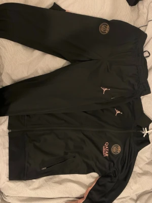 Svart PSG träningsdress Jordan - Säljer en svart träningsdress från PSG och Jordan med rosa detaljer. Setet består av zip-jacka och byxor, båda med PSG-logga och Jordan-logga samt Qatar Airways-tryck. Materialet är mjukt och stretchigt, perfekt för träning eller chill.
