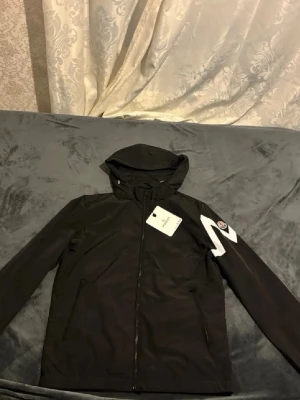 Svart vindjacka från Moncler - Snygg svart vindjacka från Moncler med huva och dragkedja framtill. Jackan har två fickor med dragkedja och en vit Moncler-logga på vänster ärm. Perfekt för dig som vill ha en stilren och sportig look.