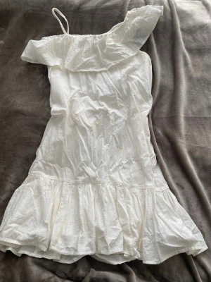 Vit offshoulder klänning Kappahl - Söt vit offshoulder klänning från Kappahl med volang upptill och nertill. Klänningen har en axelbandslös design med ett smalt axelband på ena sidan och är tillverkad i ett lätt linne tyg. Perfekt för varma dagar och sommarens alla tillfällen. 