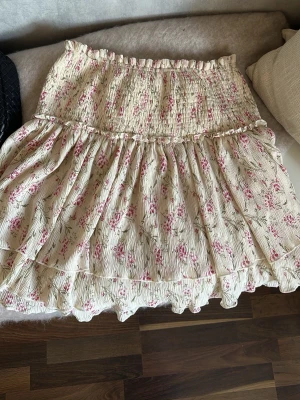 Blommig volangkjol  - Supersöt kort kjol från NA-KD i beige med små rosa blommor och volangdetaljer. Kjolen har smock i midjan för en bekväm passform och är perfekt för vår och sommar. Tillverkad i ett lätt och luftigt material. Storlek 40🌸💐