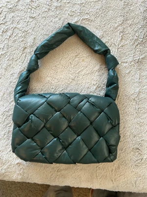 Grön handväska - Trendig handväska i mörkgrönt vadderat material med flätad design och vridet handtag. Väskan har innerfack med dragkedja och präglad logotyp på insidan. 