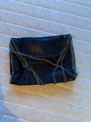 Svart axelväska med kedjedetaljer - Snygg svart axelväska i mjukt material med silverfärgade kedjor längs kanterna och som axelrem. Ungefär dataväsk storlek och kan bäras på 2 sätt och har rymliga fickor. 
