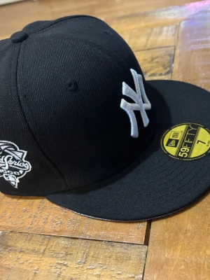 Svart New Era Yankees 59FIFTY keps - Säljer en svart New Era 59FIFTY keps med New York Yankees-logga i vitt framtill och broderad World Series-detalj på sidan. Klassisk rak skärm och broderad New Era-logga på sidan. Tillverkad i bomullstyg för en clean och sportig look.