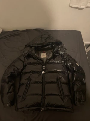 Svart dunjacka från Moncler - Snygg svart dunjacka från Moncler med glansig finish och huva. Jackan har två dragkedjeförsedda fickor framtill, Moncler-logga på ärmen och quiltad design. Perfekt för kalla vinterdagar och riktigt trendig look.