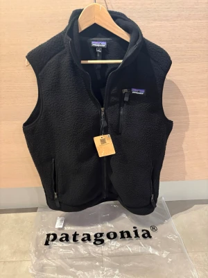 Patagonia väst - Säljer en svart fleeceväst från Patagonia med dragkedja framtill, två sidofickor med dragkedja och en bröstficka. Västen har hög krage och är gjord i mjukt, värmande fleecematerial. Perfekt för lager-på-lager och outdoor-stil.