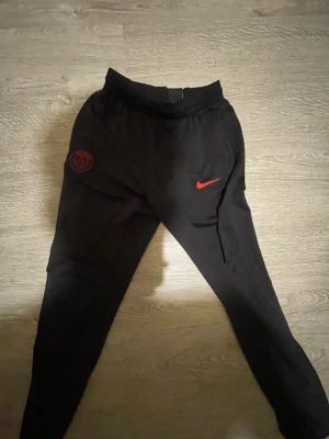 Svarta PSG mjukisbyxor från Nike - Svarta mjukisbyxor från Nike med PSG-logga och röd Nike Swoosh på ena benet. Byxorna har smal passform, resår i midjan och sidofickor. Perfekta för chill eller träning, tillverkade i mjukt material.