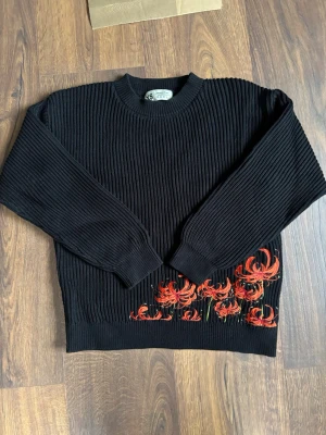 Svart stickad tröja med röda blommor - Svart ribbstickad tröja med rund halsringning och långa ärmar. Nedtill finns broderade röda och orange blommor med gröna detaljer både fram och bak. Perfekt för dig som vill ha en unik och färgstark detalj i garderoben.