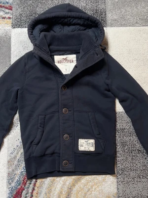 Mörkblå hoodie från Hollister - Snygg mörkblå hoodie från Hollister med knappar framtill, två fickor och en stor huva. Hoodien har ribbad krage och en patch med Hollister-logga på ena fickan. Tillverkad i mjukt bomullsmaterial, perfekt för en avslappnad stil.