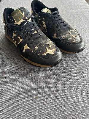 Valentino Rockrunners - Snygga sneakers från Valentino med svart och beige camomönster och guldiga stjärnor. Skorna har svarta snören, beige detaljer och nitar på hälen. Materialmix av läder och textil. Perfekta för dig som vill sticka ut med en unik design.Orginal Box medföljs Ej. 