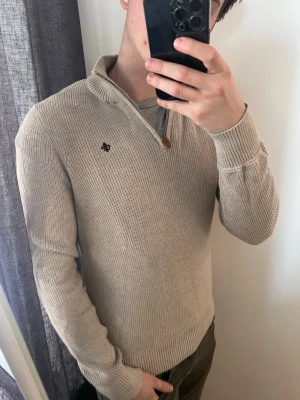 Beige stickad tröja från Morris - Snygg beige stickad tröja från Morris med halv dragkedja och ribbad krage. Tröjan har en liten mörkblå logga broderad på bröstet och ribbade muddar vid ärmslut och nederkant. Perfekt för lager på lager och stilren look. Storlek S