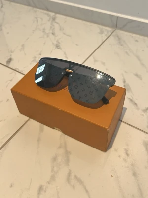 Louis Vuitton svarta shield-solglasögon - Säljer ett par svarta shield-solglasögon från Louis Vuitton med spegelglas och ikoniskt LV-monogram över hela linsen. Glasögonen har en futuristisk, kantig form och bred båge. Perfekta för dig som vill sticka ut med en lyxig accessoar.