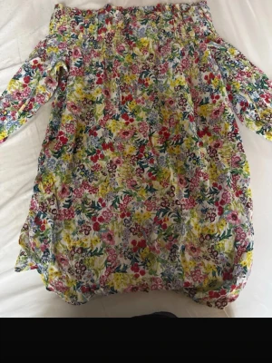 Blommig offshoulder klänning - Färgglad offshoulder klänning med blommigt mönster i rött, gult, blått och grönt. Klänningen har trekvartsärm och en lös passform, perfekt för varma dagar. Tillverkad i ett lätt och luftigt material.