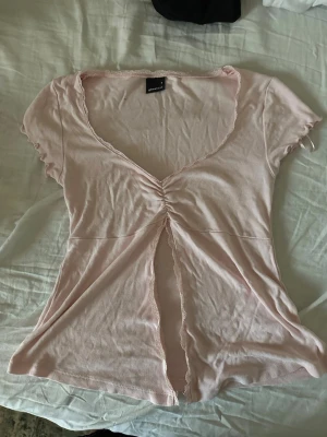 Ljusrosa omlottopp med spetsdetalj - Söt ljusrosa omlottopp från Monki med korta ärmar och v-ringning. Toppen har en snygg rynkning framtill, spetskant längs öppningen och en liten slits. Perfekt för dig som gillar feminina detaljer och mjuka material.