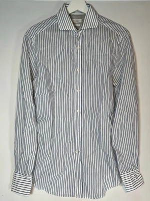 BRUNELLO CUCINELLI - Klassisk vit skjorta med tunna blå ränder. Skjortan har lång ärm, knappar framtill och krage. Tillverkad i bomull för en skön och luftig känsla. Perfekt för en clean och stilren look.