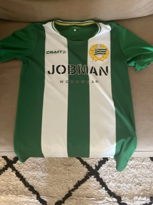 Hammarby matchtröja Craft grön/vit - Säljer en officiell Hammarby matchtröja från Craft i grönt och vitt med klubbmärke på bröstet och JOBMAN Workwear som sponsortryck. Tröjan har korta ärmar, rund hals och är tillverkad i ett lätt, funktionellt material perfekt för fotboll eller träning.