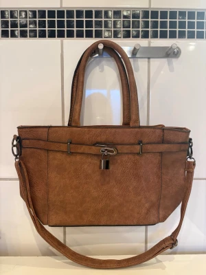 Brun handväska med axelrem - Snygg brun handväska i konstläder med både handtag och justerbar axelrem. Väskan har silverfärgade metalldetaljer och ett dekorativt lås framtill. Stilren och rymlig modell som passar till många olika outfits.