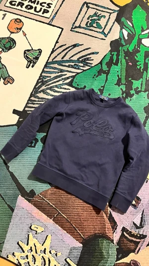 Mörkblå stickad tröja Polo Ralph Lauren - Mörkblå stickad tröja från Polo Ralph Lauren med broderad logga framtill. Tröjan har rund halsringning och ribbade muddar vid ärmslut och nederkant. Perfekt för en avslappnad och stilren look.