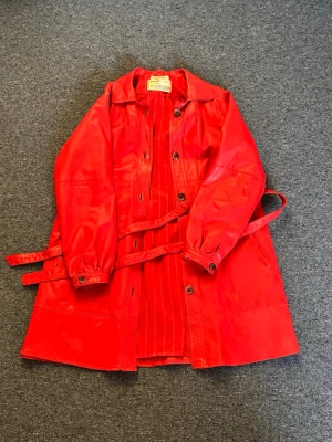Vintage röd skinnjacka från 60 talet - Vintage röd skinnjacka från 60 talet med knappar framtill och tillhörande bälte i midjan. Jackan har en klassisk krage, långa ärmar med knappdetaljer vid ärmslut och en längre, rak passform. Jackan är från Wasa päls gjord av Torsten Albertsson. Perfekt för dig som vill sticka ut med färg. Eller bara erhålla en vintage look. Den är använd men i mycket bra skick. Pris kan diskuteras. 
