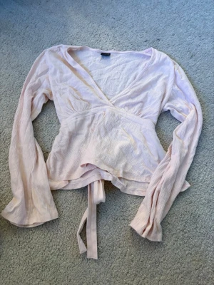 Ljusrosa omlottblus med knytband - Supersöt ljusrosa tröja med långa ärmar och knytband i midjan. Tröjan har en lätt skrynklig struktur och v-ringad halsringning. Perfekt att styla med jeans eller kjol. Tröjan är från Gina trickot och har aldrig använt den.