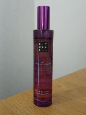 Rituals The Ritual of Yozakura mist - En elegant hår- och kroppsmist från Rituals, The Ritual of Yozakura. Flaskan är cylinderformad i lila och rosa glas med en blank lila kork. Doften är inspirerad av körsbärsblom och har en fräsch, blommig ton. Passar perfekt för dig som vill ha en lätt och fräsch parfym. Ca 90% full.