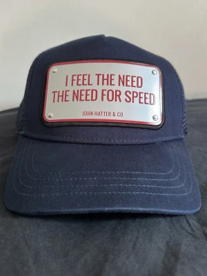 Marinblå keps John Hatter & Co - Snygg marinblå keps från John Hatter & Co med ett metallmärke framtill där det står 'I FEEL THE NEED THE NEED FOR SPEED' i röd text. Klassisk trucker-modell med mesh baktill och justerbar rem. Perfekt för dig som gillar statement-plagg och coola accessoarer. 