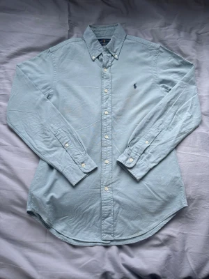 Ljusblå skjorta från Ralph Lauren - Klassisk ljusblå skjorta från Ralph Lauren i slim fit-modell. Skjortan har långa ärmar, knappar framtill och den ikoniska broderade loggan på bröstet. Tillverkad i mjuk bomull och passar perfekt till både jeans och chinos.