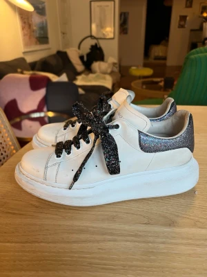 Alexander McQueen sneakers - Chunky vita sneakers från Alexander McQueen med glittriga svarta skosnören och glitterdetalj på hälen. Klassisk låg modell med bred sula och rundad tå. Perfekta för dig som vill sticka ut lite extra med dina skor.                                                          Inköpts 2025 så finns lite slitage på hälen, där lite glitter har skavts. Inköpta i New York för 5500kr. 
