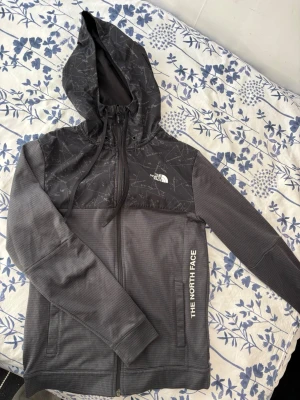 Grå hoodie från The North Face - Snygg grå hoodie från The North Face med svart mönstrad huva och axelparti. Dragkedja framtill, två fickor och justerbar huva med snören. Logga på bröstet och längs sidan. Tillverkad i mjukt, strukturerat material som känns bekvämt och sportigt.