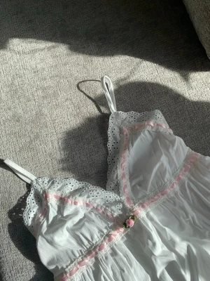 Romantiskt spetslinne  - Söt vit linnetopp med tunna axelband, spetskant och broderade blommor längs kanterna. Toppen har rosa banddetaljer och en liten ros i mitten framtill. Perfekt för en romantisk och feminin stil 🌸