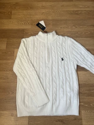 Vit kabelstickad tröja Polo Ralph Lauren - Kabelstickad vit tröja från Polo Ralph Lauren med halv dragkedja och hög krage. Tröjan har det klassiska mörkblå logobroderiet på bröstet och ribbade muddar vid ärmslut och nederkant. Perfekt för lager på lager och en clean look.
