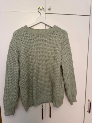 Ljusgrön stickad tröja från 157 - Mysig ljusgrön stickad tröja från 157 med grovstickat mönster och rund hals. Tröjan har långa ärmar och ribbade muddar vid ärmslut och nederkant. Perfekt för kalla dagar och enkel att matcha med jeans eller kjol.